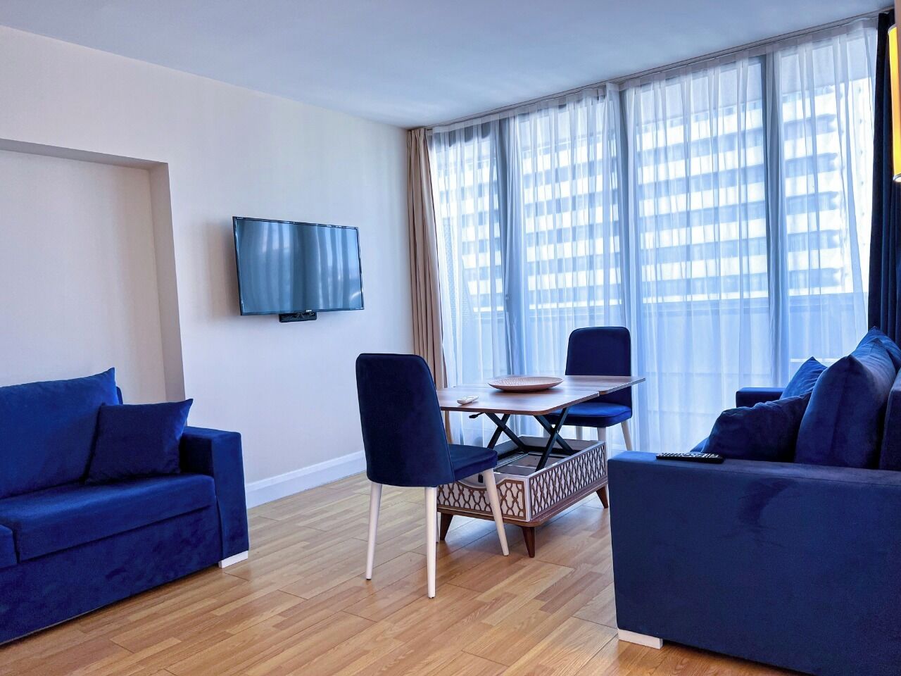 A modern living room with a sofa, a dining table with chairs, a TV mounted on the wall, and large windows with sheer curtains.