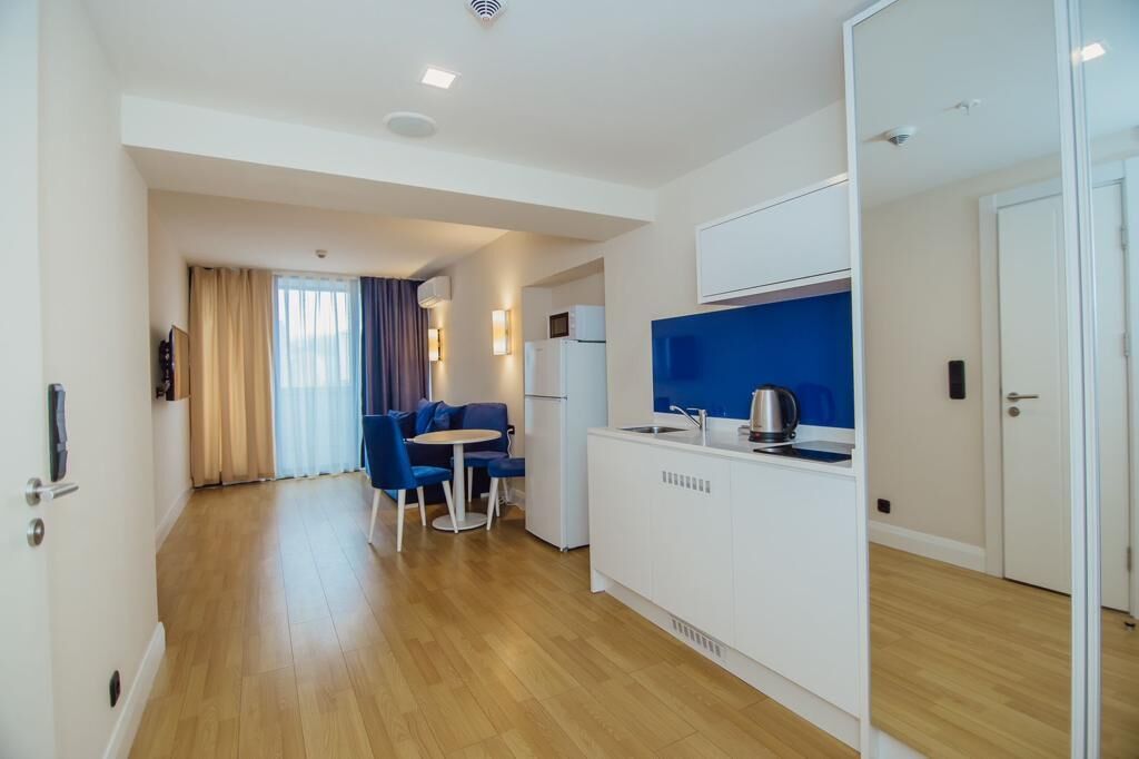 A modern hotel room with a wooden floor, a kitchenette, a dining area with a table and chairs, and a large window with curtains.