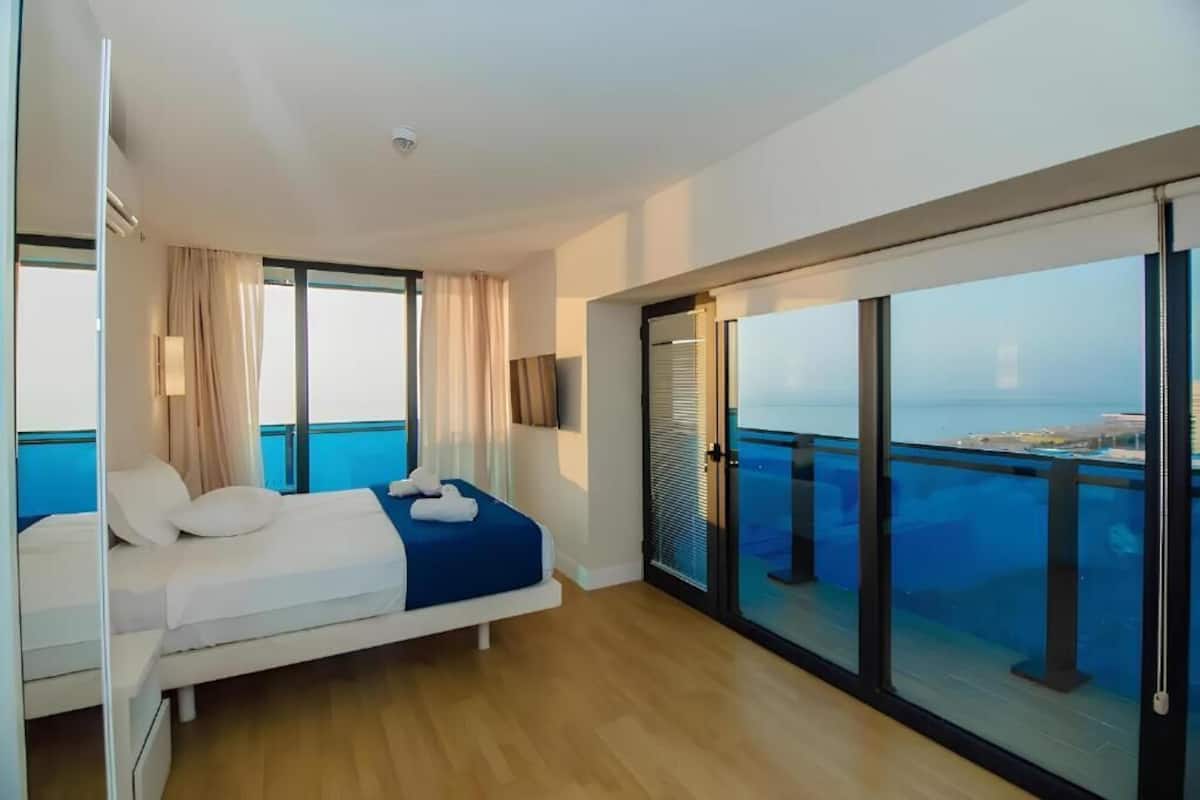 A modern hotel room with a large bed, a balcony with a city view, and a sliding glass door.