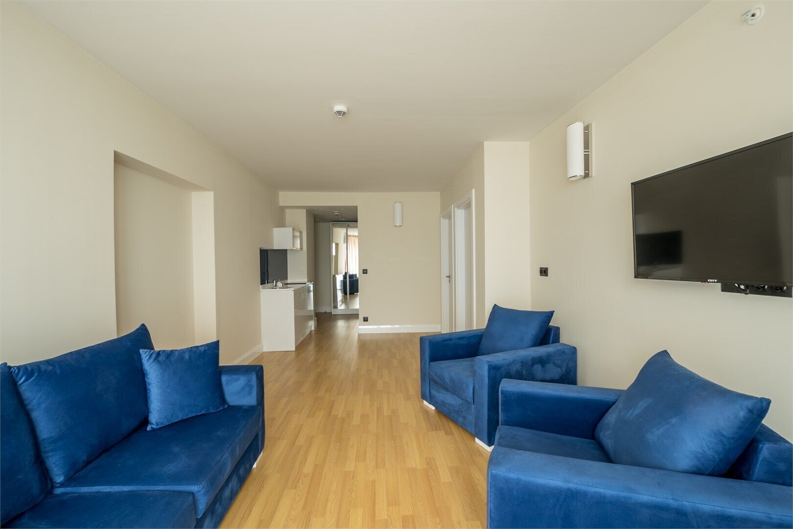 A hallway with a TV mounted on the wall, a kitchen area, and a seating area with blue sofas.
