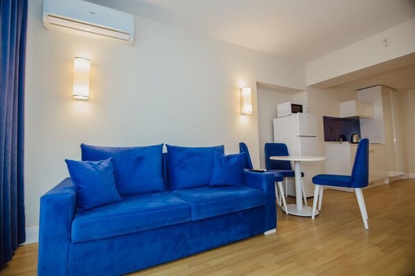 Un salon moderne avec un canapé bleu, une petite table à manger et des chaises. On aperçoit à l’arrière-plan un coin cuisine avec des meubles blancs et un four à micro-ondes.