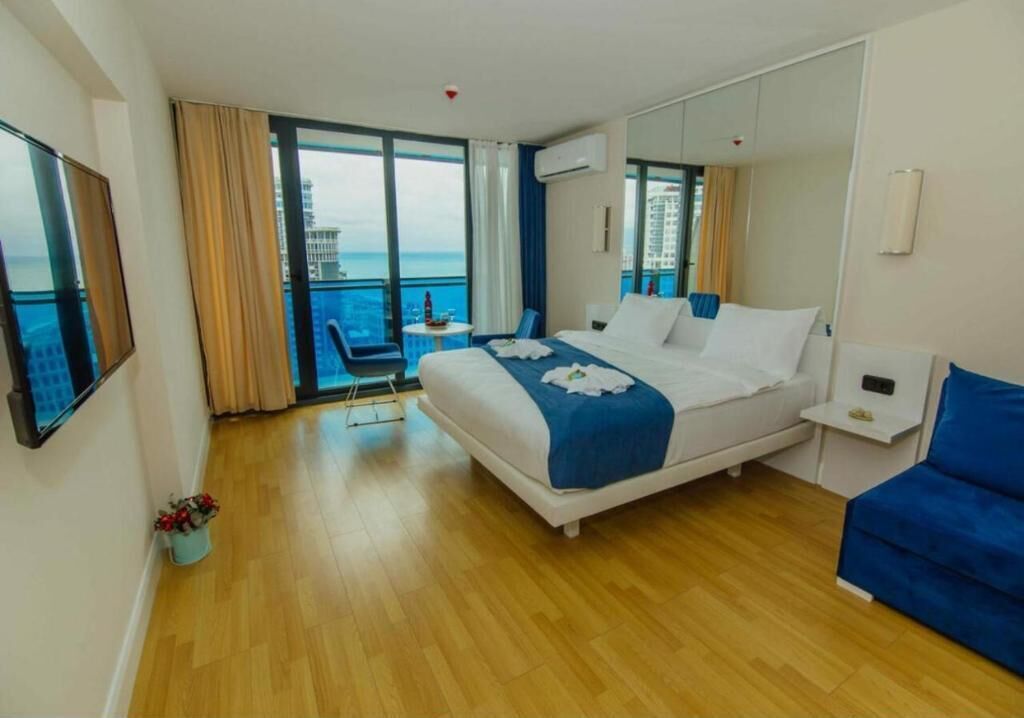 Habitación de hotel con una cama grande, un sofá azul, una pequeña mesa con una bebida, una silla y vistas a la ciudad y al mar a través de amplias ventanas.