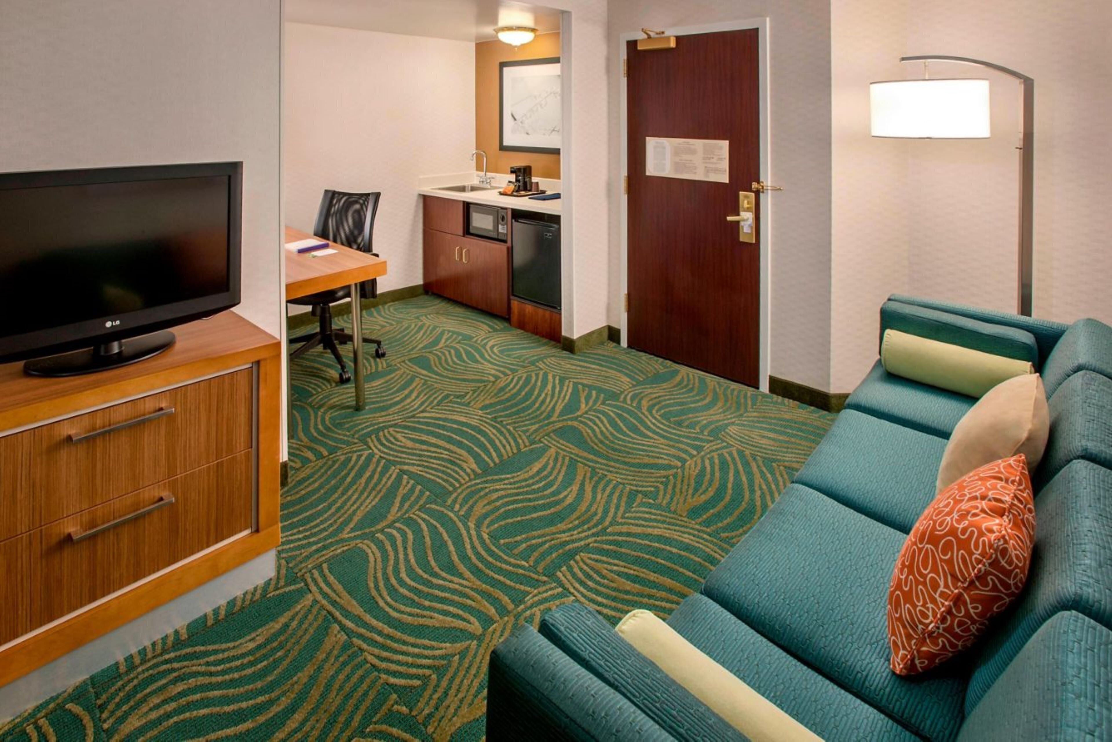 A hotel room with a television, a kitchenette, a sofa, and a patterned carpet.