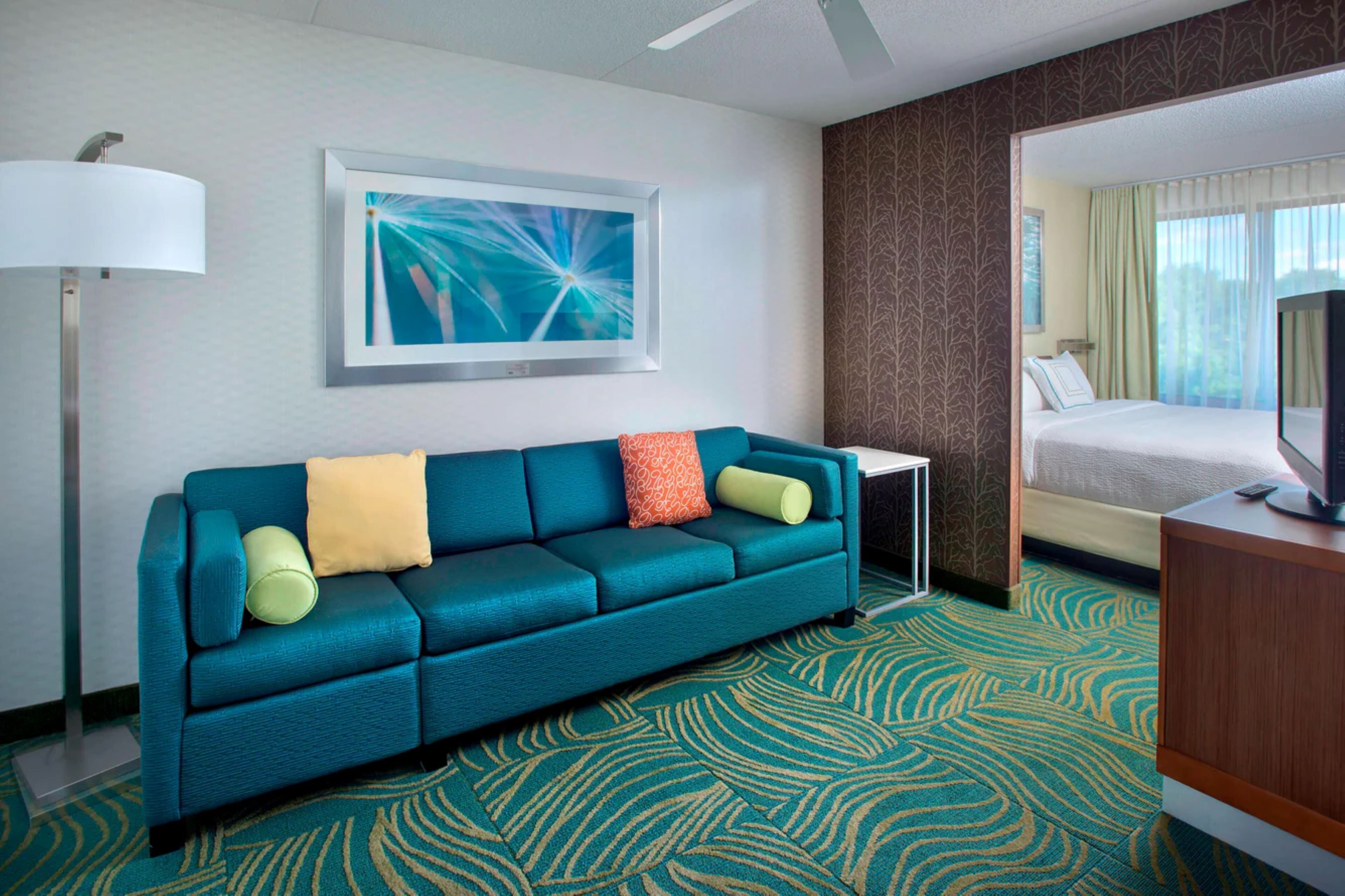 A hotel room with a teal sofa, a framed abstract painting, a television, and a patterned carpet.
