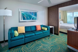 Habitación de hotel con un sofá color turquesa, un cuadro abstracto enmarcado, un televisor y una alfombra estampada.