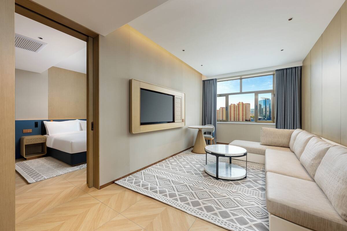 A modern hotel room with a bed, a sofa, a TV, and a city view.