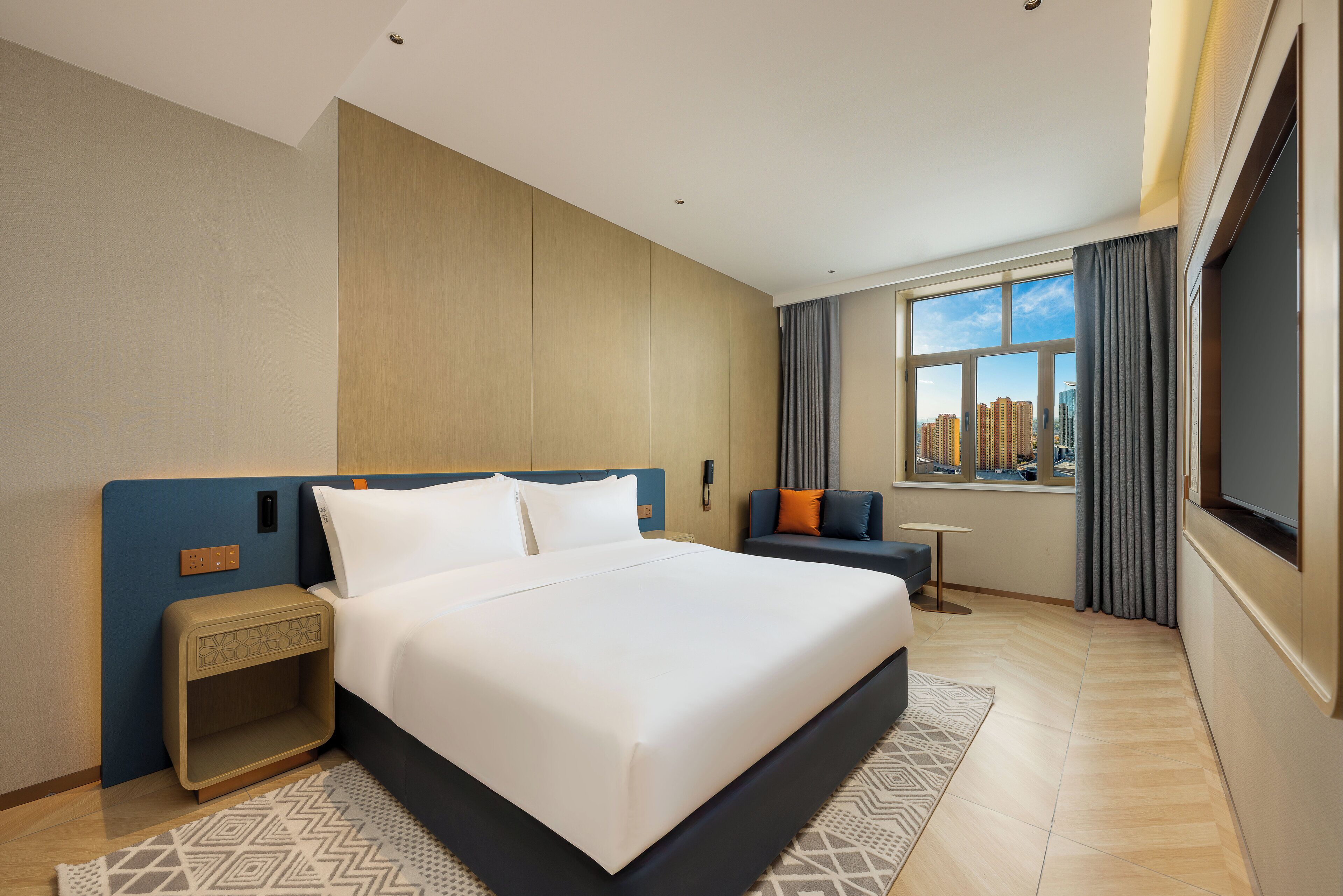 A modern hotel room with a large bed, a small side table, a sofa, a TV, and a city view.
