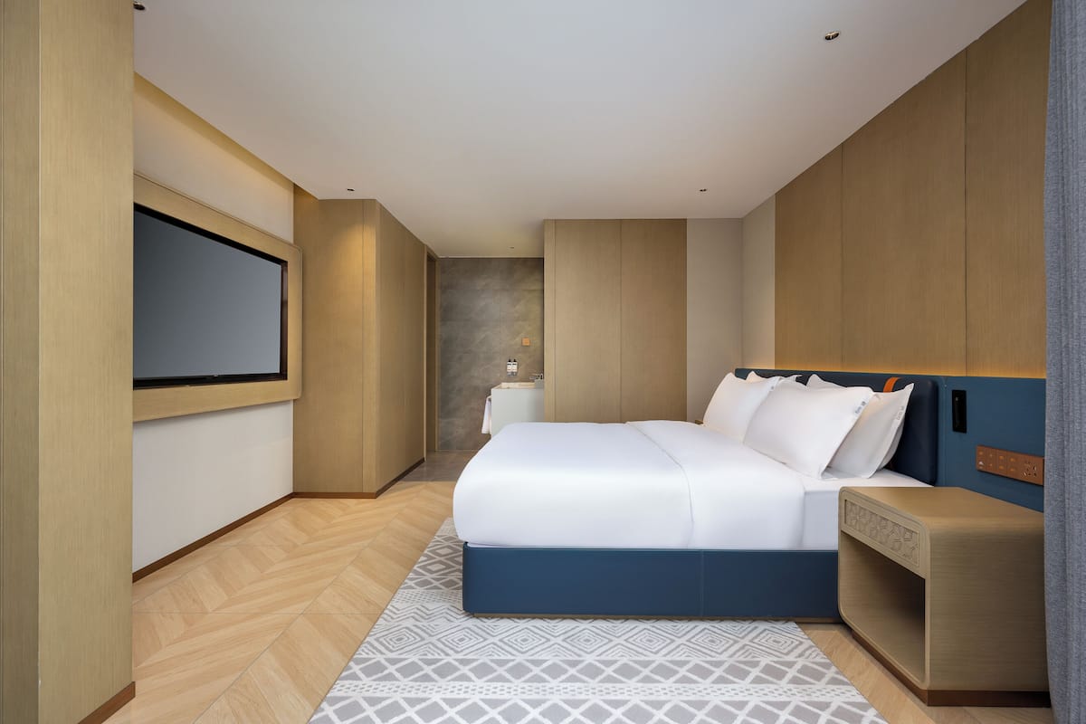 A modern hotel room with a large bed, a bedside table, a TV mounted on the wall, and a small bedside lamp.