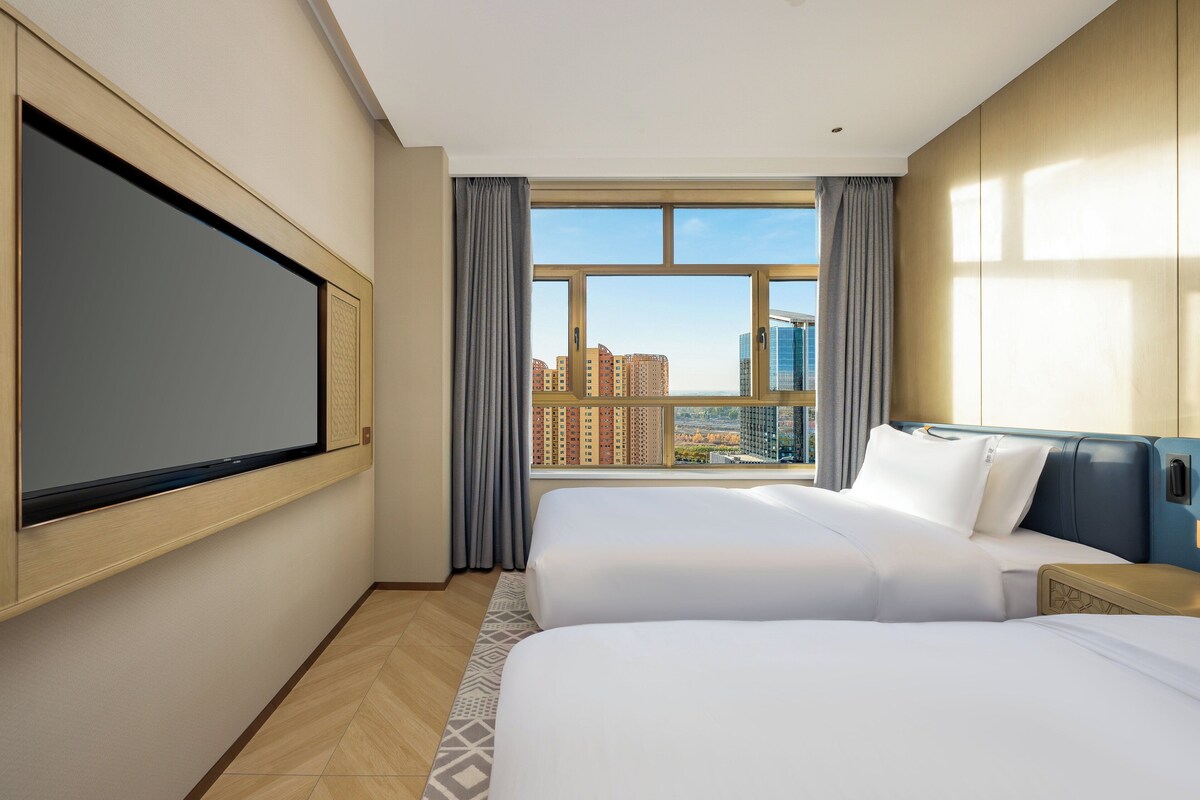 Habitación de hotel con dos camas, un ventanal grande con vistas a la ciudad, un televisor de pantalla plana en la pared y un cabecero de madera.