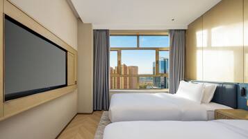 A hotel room with two beds, a large window with a city view, a flat-screen TV mounted on the wall, and a wooden headboard.