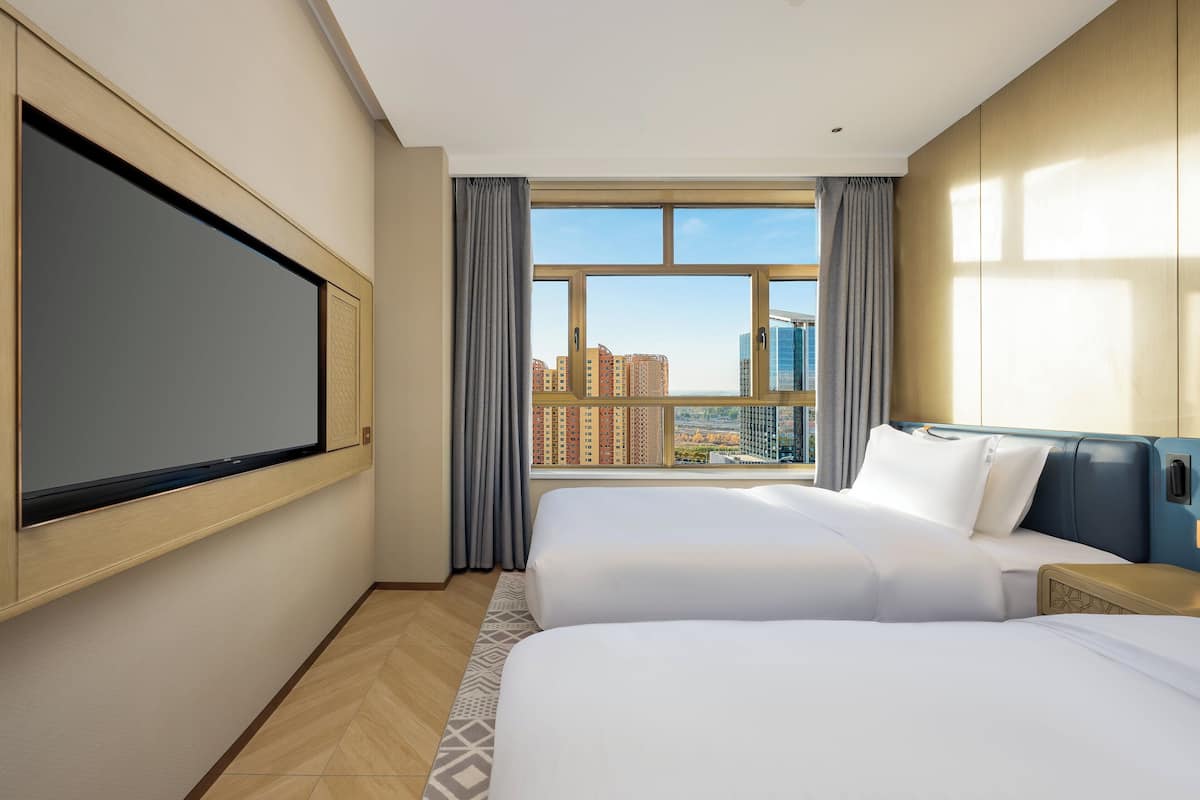 A hotel room with two beds, a large window with a city view, a flat-screen TV mounted on the wall, and a wooden headboard.