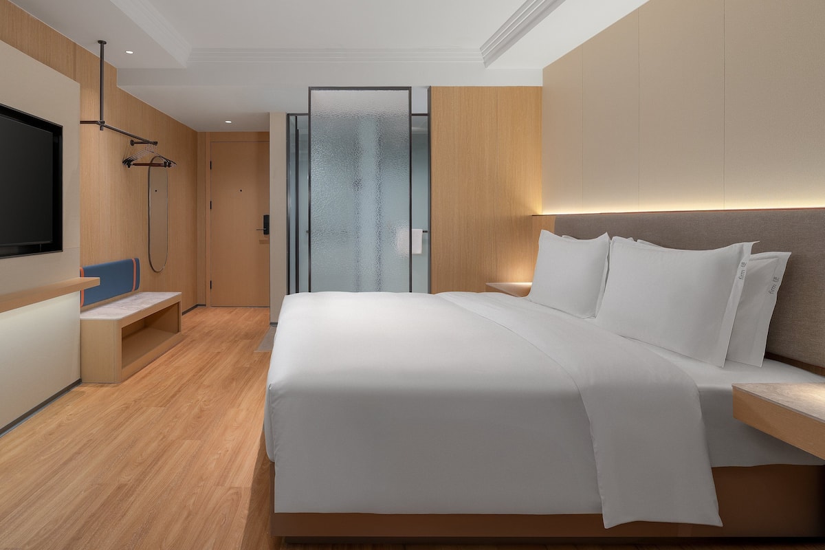A modern hotel room with a large bed, a TV mounted on the wall, a seating area with a bench, and a glass partition.