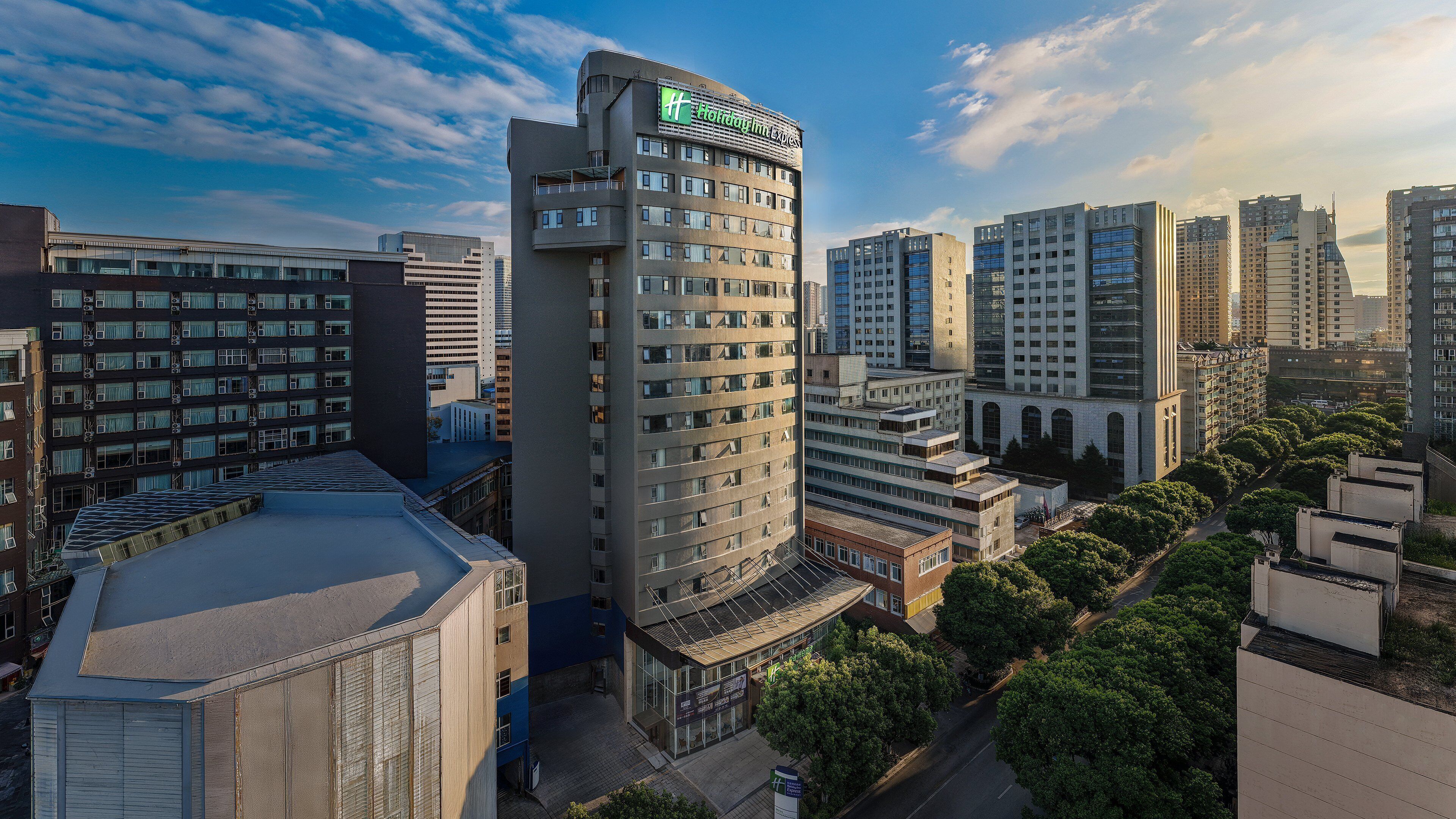 A modern hotel with a prominent sign, surrounded by high-rise buildings and greenery.