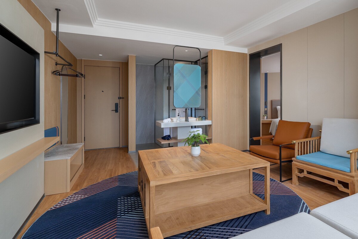 A modern hotel room with a wooden table, a sofa, a chair, a TV, and a bathroom with a mirror and sink.