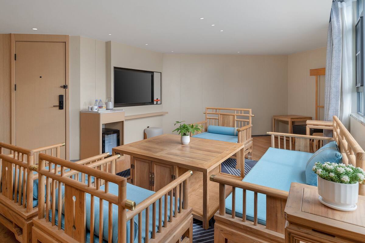 A modern, well-lit room with a wooden dining table, blue cushioned seating, a TV mounted on the wall, and a window with curtains.