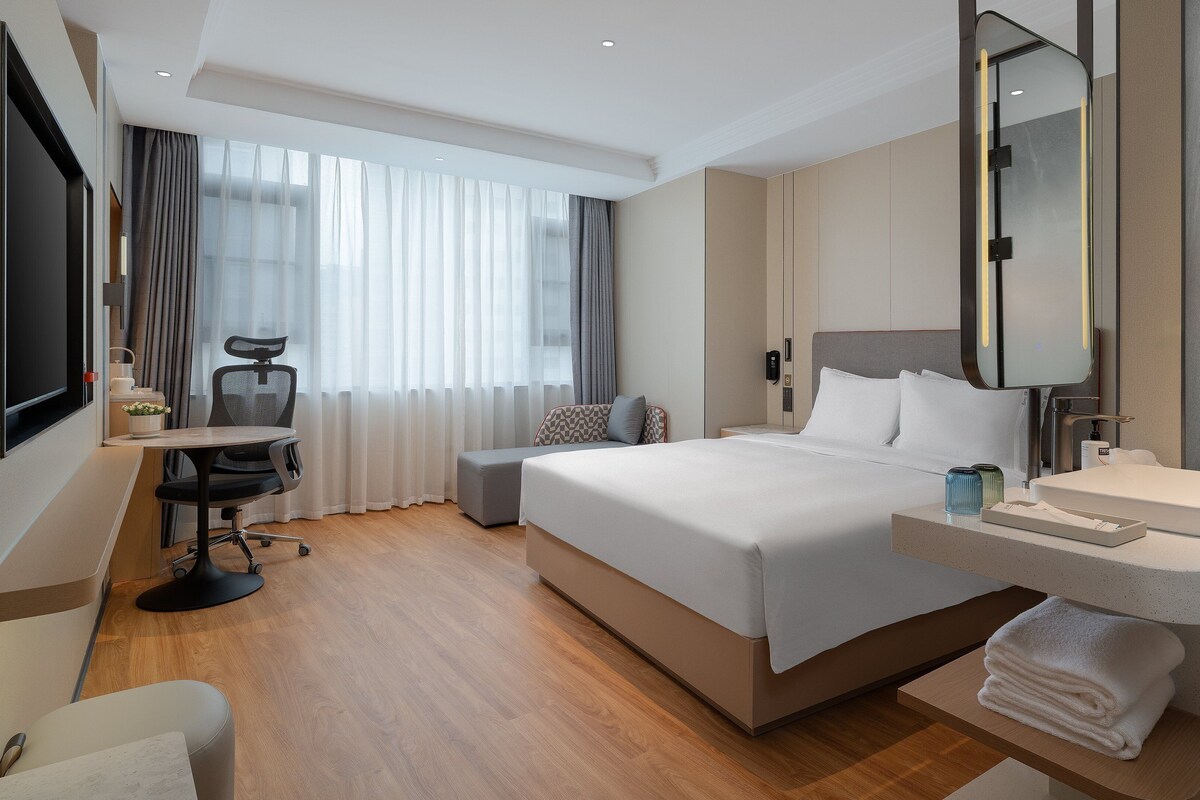 A hotel room with a large bed, a desk with a chair, a TV, and a bathroom with a mirror and sink.