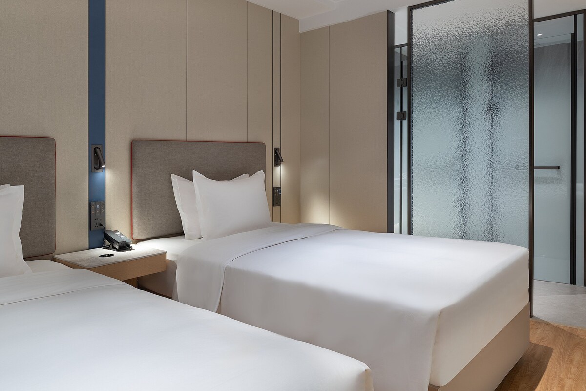 A modern hotel room with two beds, a glass shower enclosure, and a wooden floor.