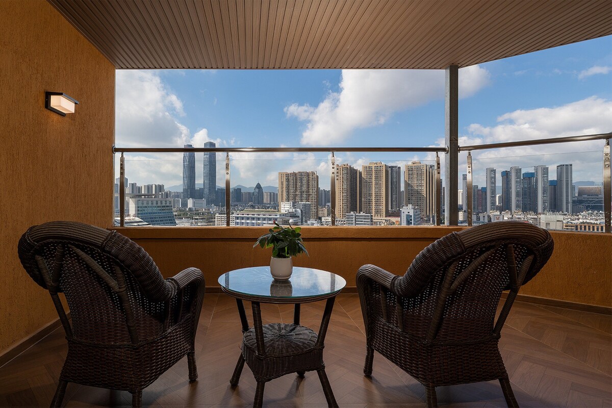 A balcony with wicker chairs, a glass table, and a potted plant. A cityscape is visible through large windows.