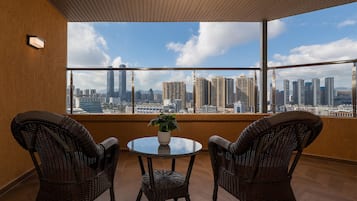 Un balcon avec des fauteuils en osier, une table en verre et un pot de fleurs. On peut apercevoir le paysage urbain à travers de grandes fenêtres.