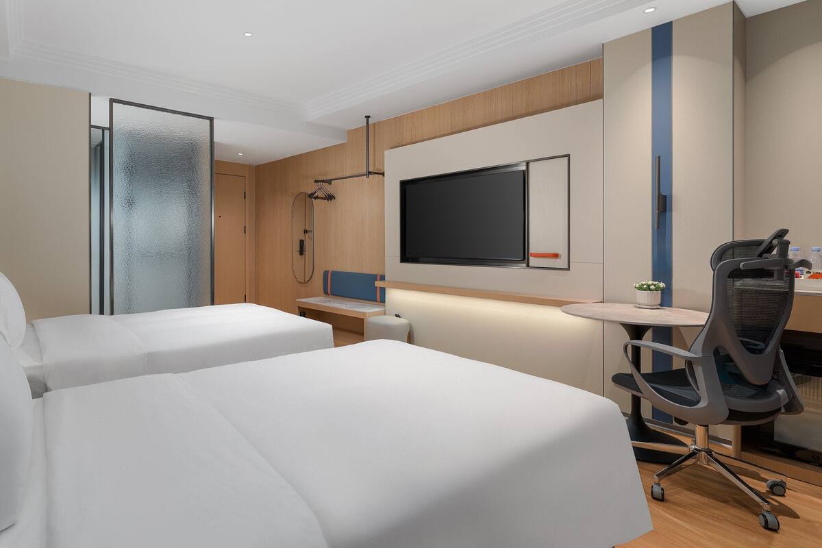 A hotel room with two beds, a desk, a chair, a TV, and a shower area.