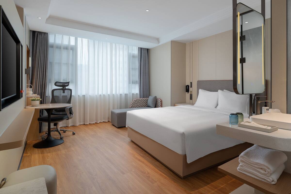 A hotel room with a large bed, a desk with a chair, a TV, and a bathroom with a mirror and sink.