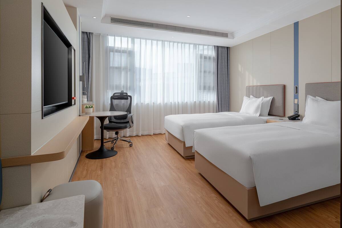 A hotel room with two beds, a desk with a chair, a TV, and a large window with curtains.