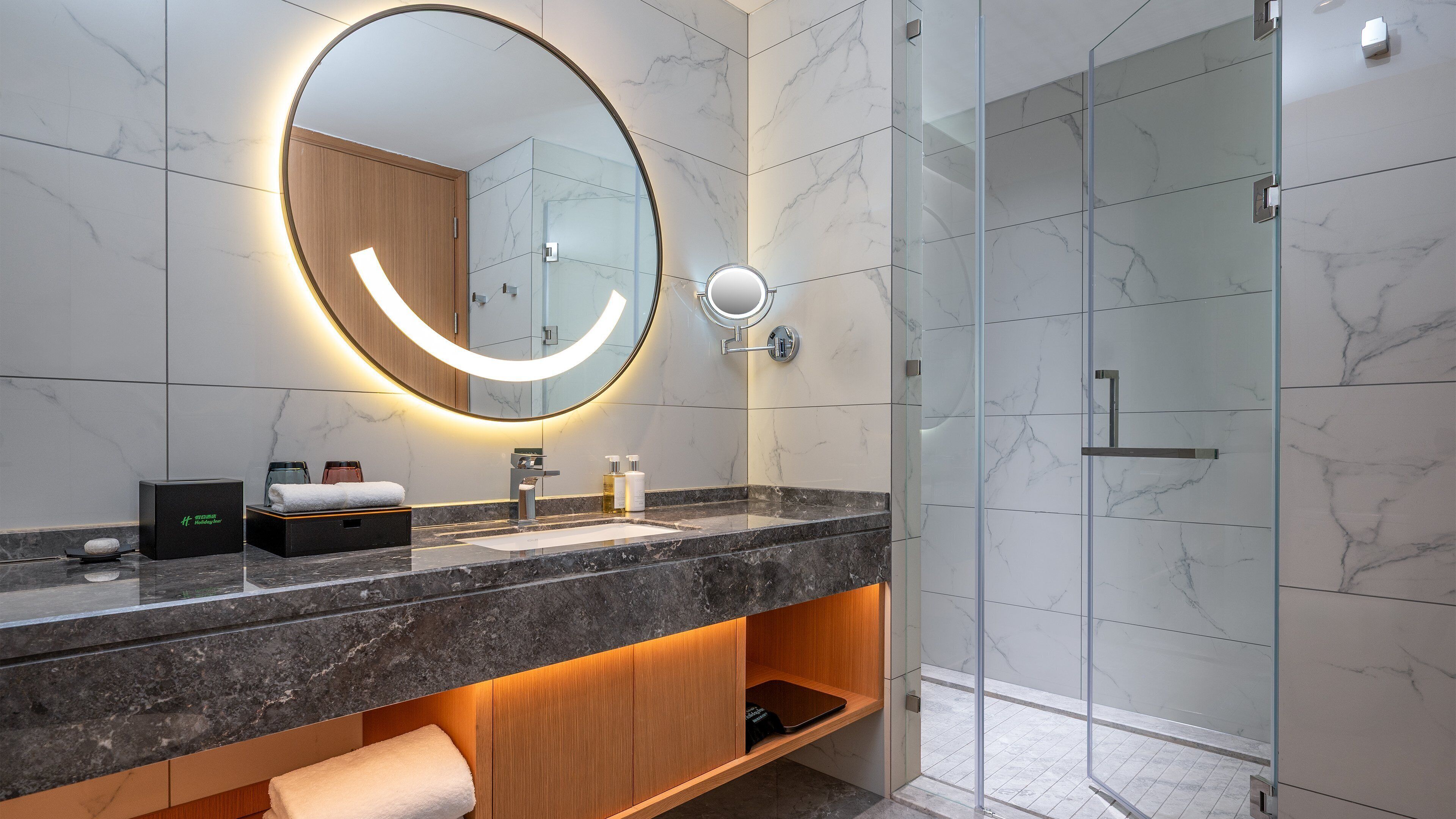 Un baño moderno con tocador de mármol, un espejo grande con iluminación integrada, cabina de ducha de vidrio y estante de madera con toallas.