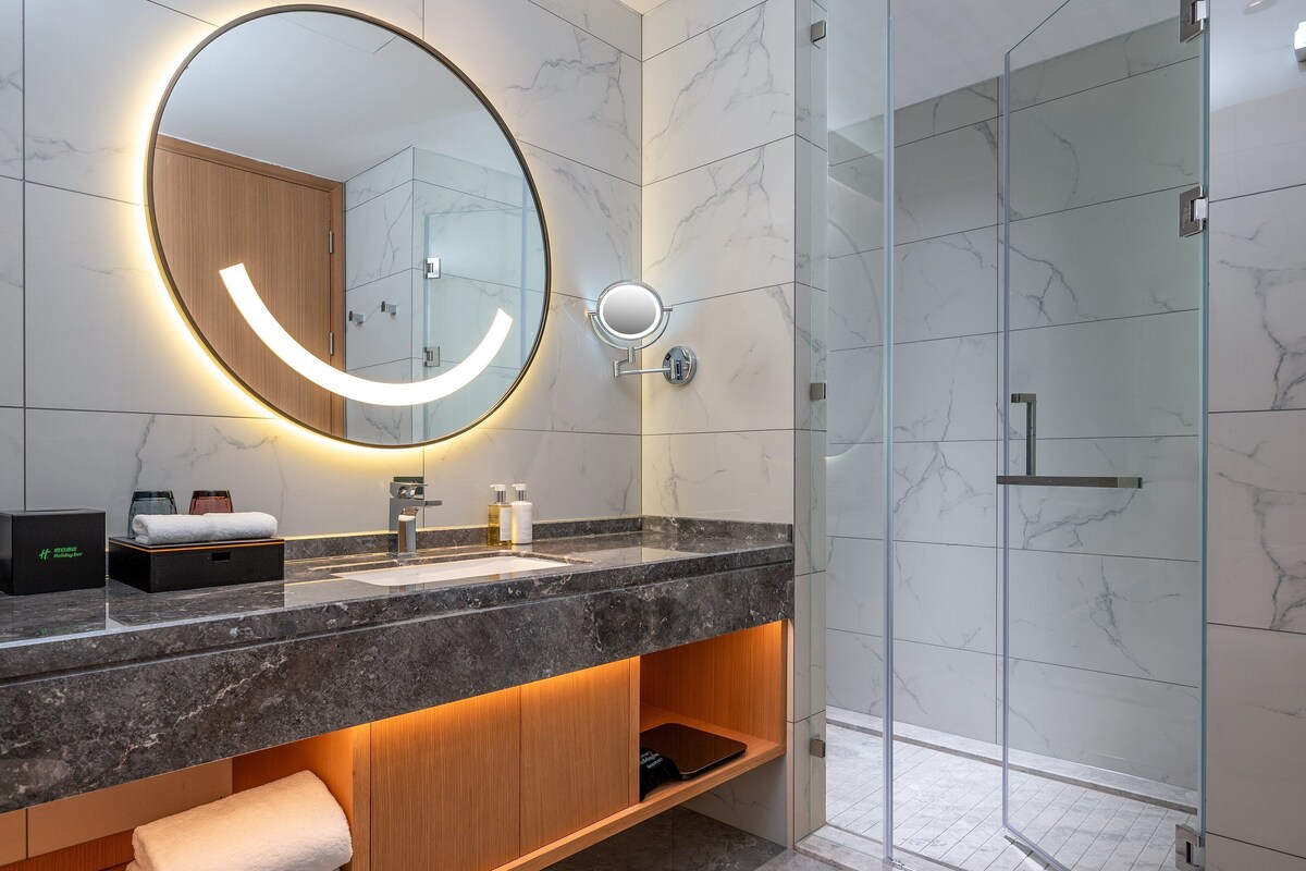 Une salle de bain moderne avec un meuble-lavabo en marbre, un grand miroir doté d’un éclairage intégré, une cabine de douche en verre et une étagère en bois où sont posées des serviettes.