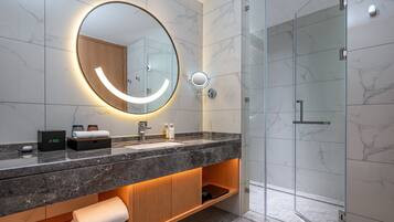 Un baño moderno con tocador de mármol, un espejo grande con iluminación integrada, cabina de ducha de vidrio y estante de madera con toallas.