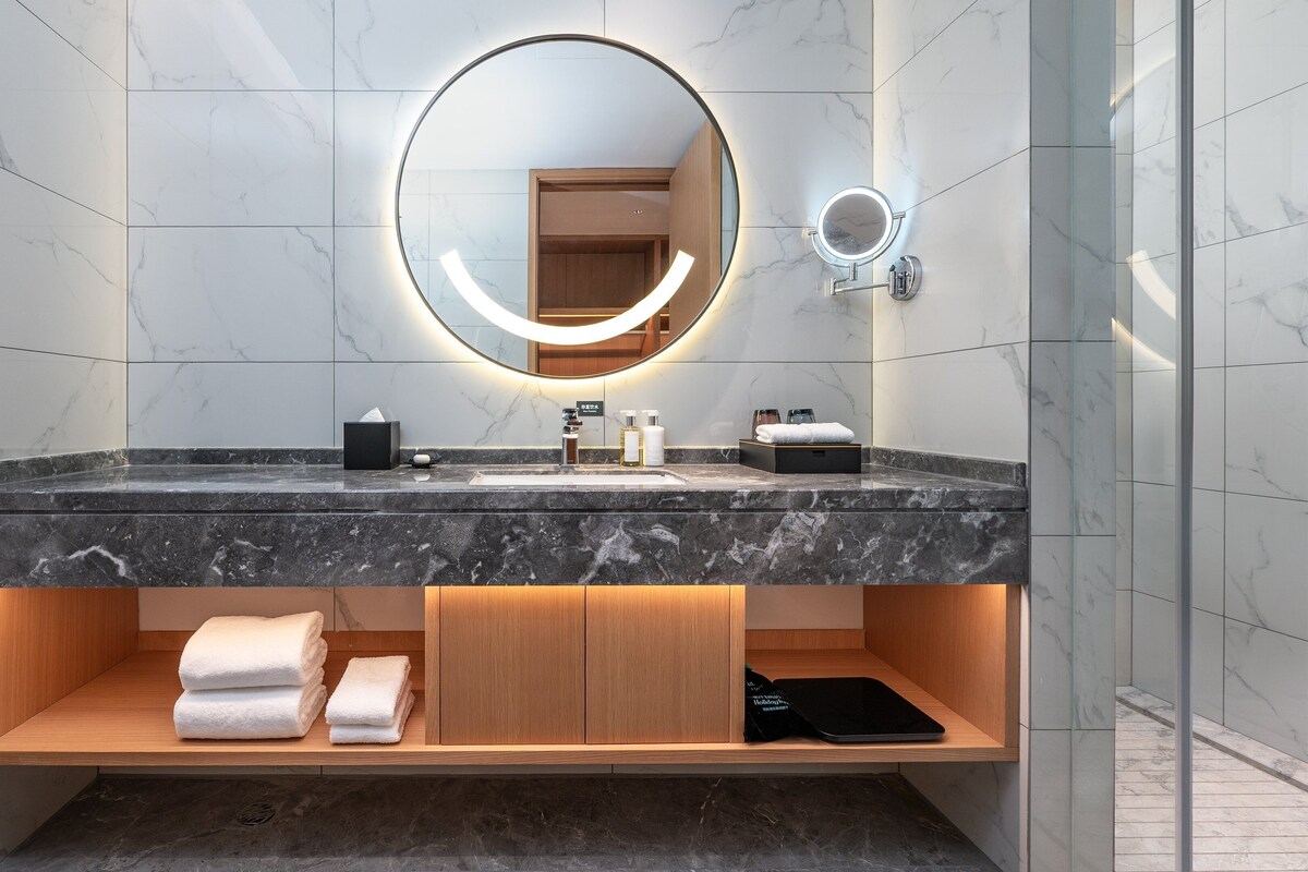 Une salle de bain moderne avec un meuble-lavabo en marbre, un grand miroir rond doté d’un éclairage intégré et une étagère en bois où sont posées des serviettes.
