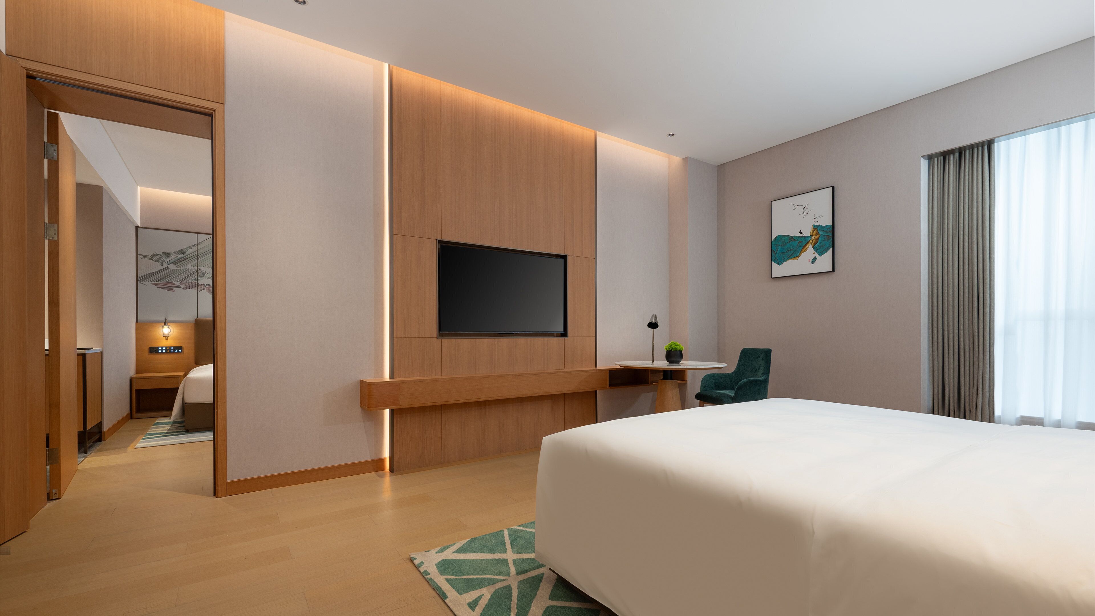A modern hotel room with a large bed, a flat-screen TV mounted on the wall, a small table with a lamp, and a chair.