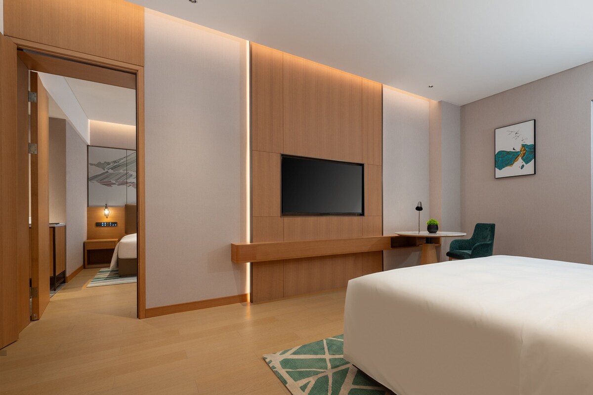 Une chambre d’hôtel moderne avec un grand lit, un téléviseur à écran plat fixé au mur, une petite table avec une lampe et une chaise.