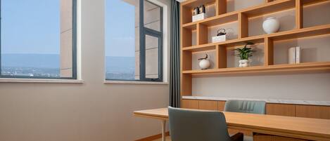 A room with a large window, a wooden desk, a chair, and a built-in shelving unit with decorative items.