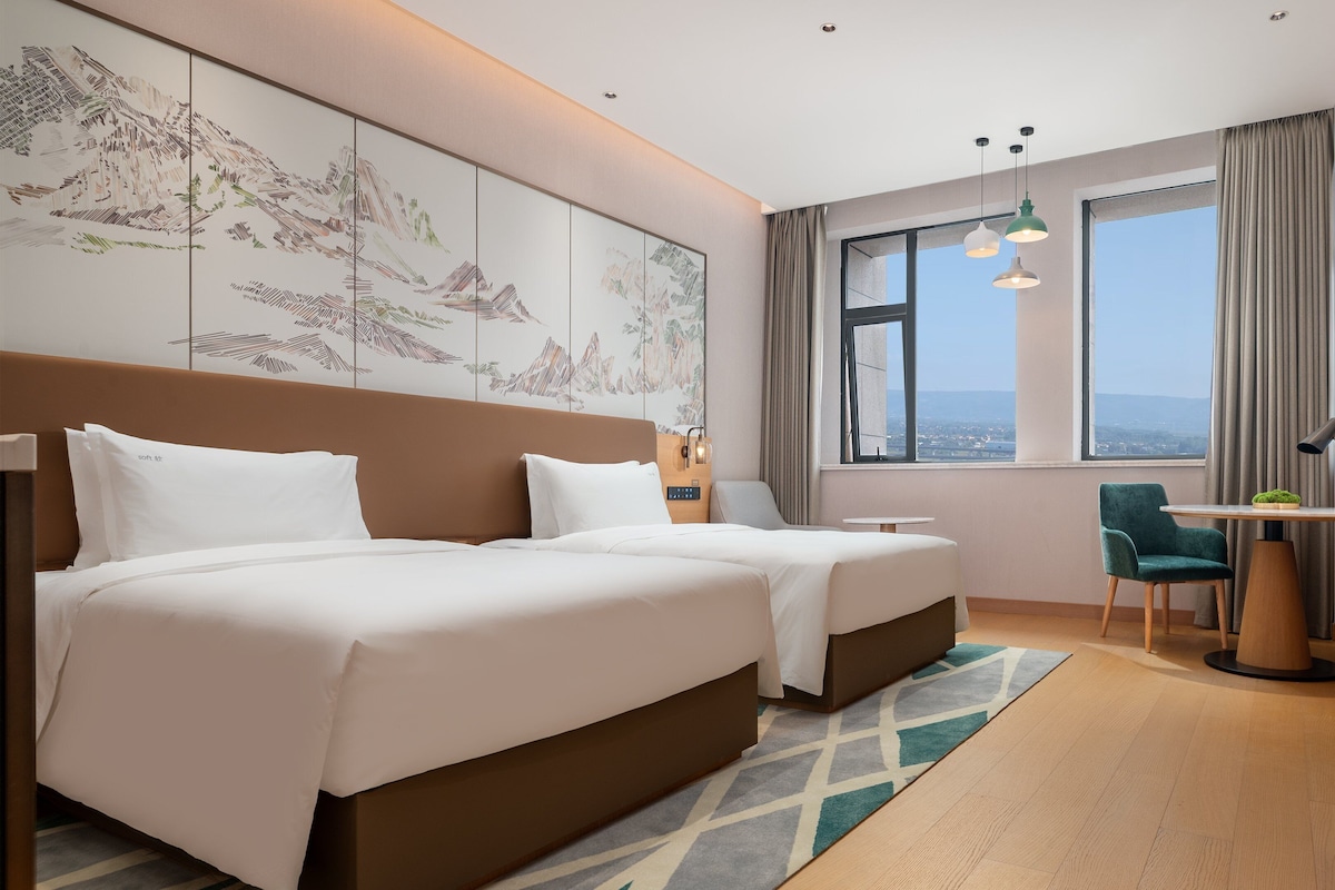 Habitación de hotel con dos camas, un escritorio y una silla, un ventanal con vistas, y obras de arte en la pared.