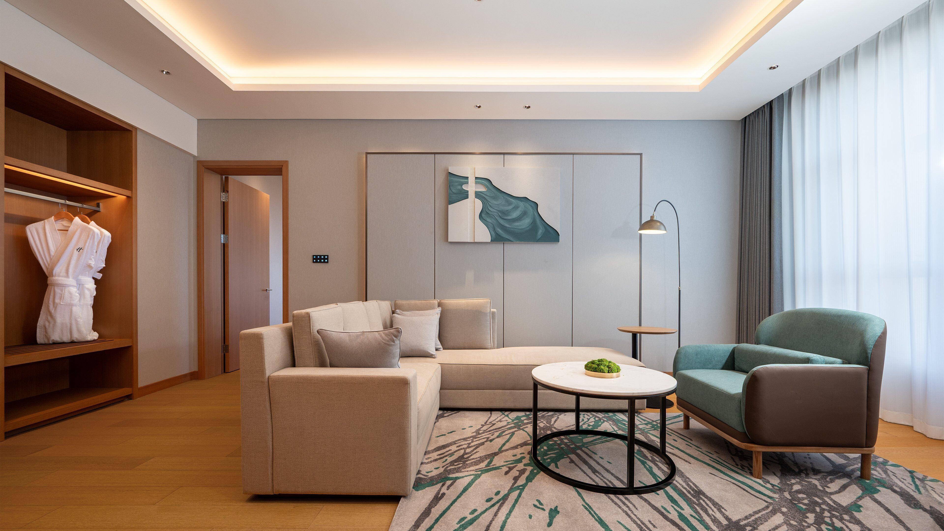 Una habitación de hotel moderna con sofá, sillón y una pequeña mesa con una planta. Se ve un armario con batas colgadas en su interior, y un cuadro en la pared.