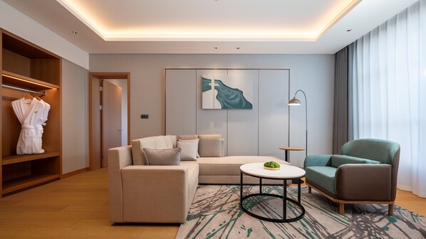 Quarto de hotel moderno com sofá, poltrona e uma pequena mesa com uma planta. Possui um guarda-roupa com roupões pendurados no interior, e uma pintura na parede.