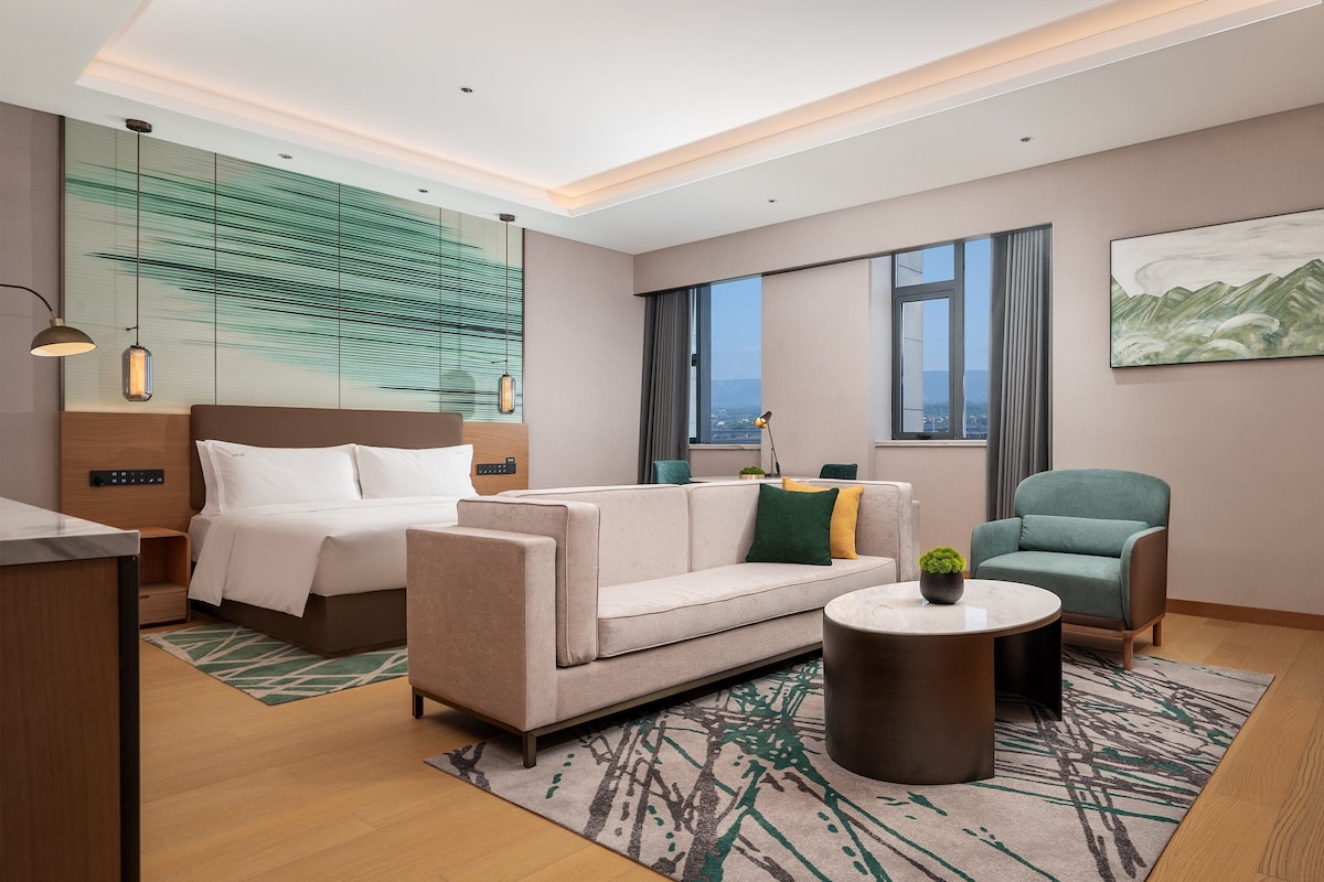 Habitación de hotel moderna con cama, sofá, sillón y vista de la ciudad a través de amplios ventanales.