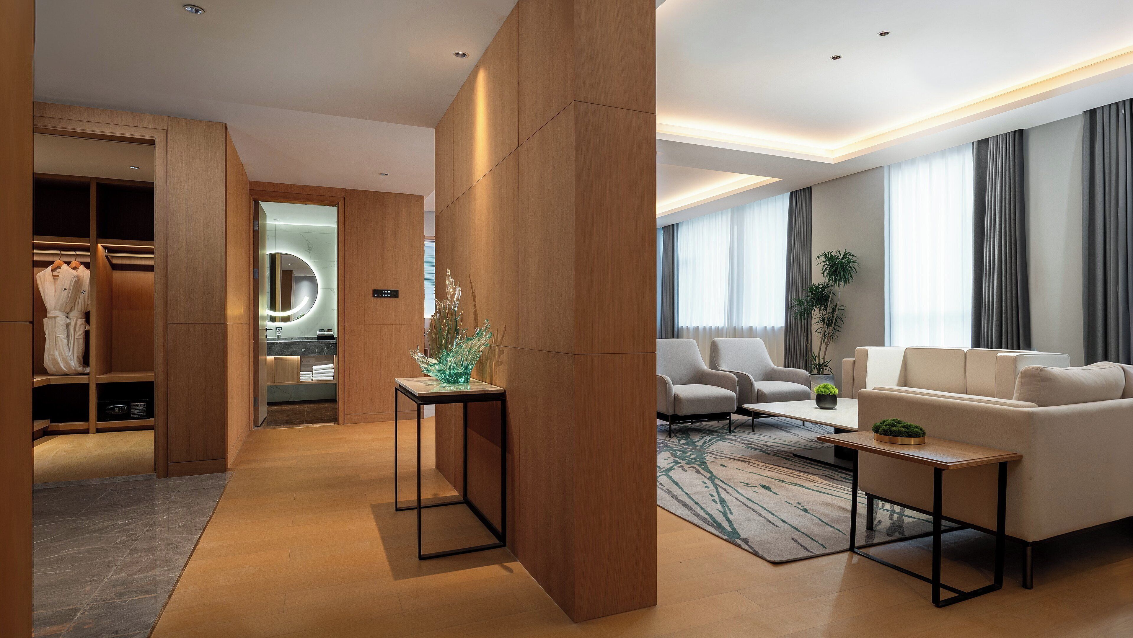 Una habitación de hotel moderna con un armario de madera, una zona de estar con sofá y sillones, una mesita con una planta y un espejo grande con luz circular.