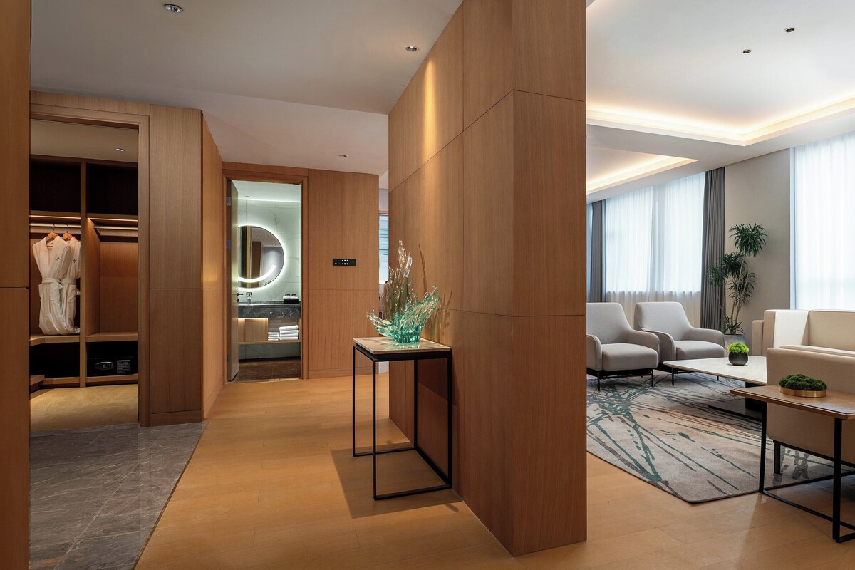 Une chambre d’hôtel moderne avec une armoire en bois, un coin salon comprenant un canapé et des fauteuils, une petite table avec une plante et un grand miroir avec un éclairage circulaire.