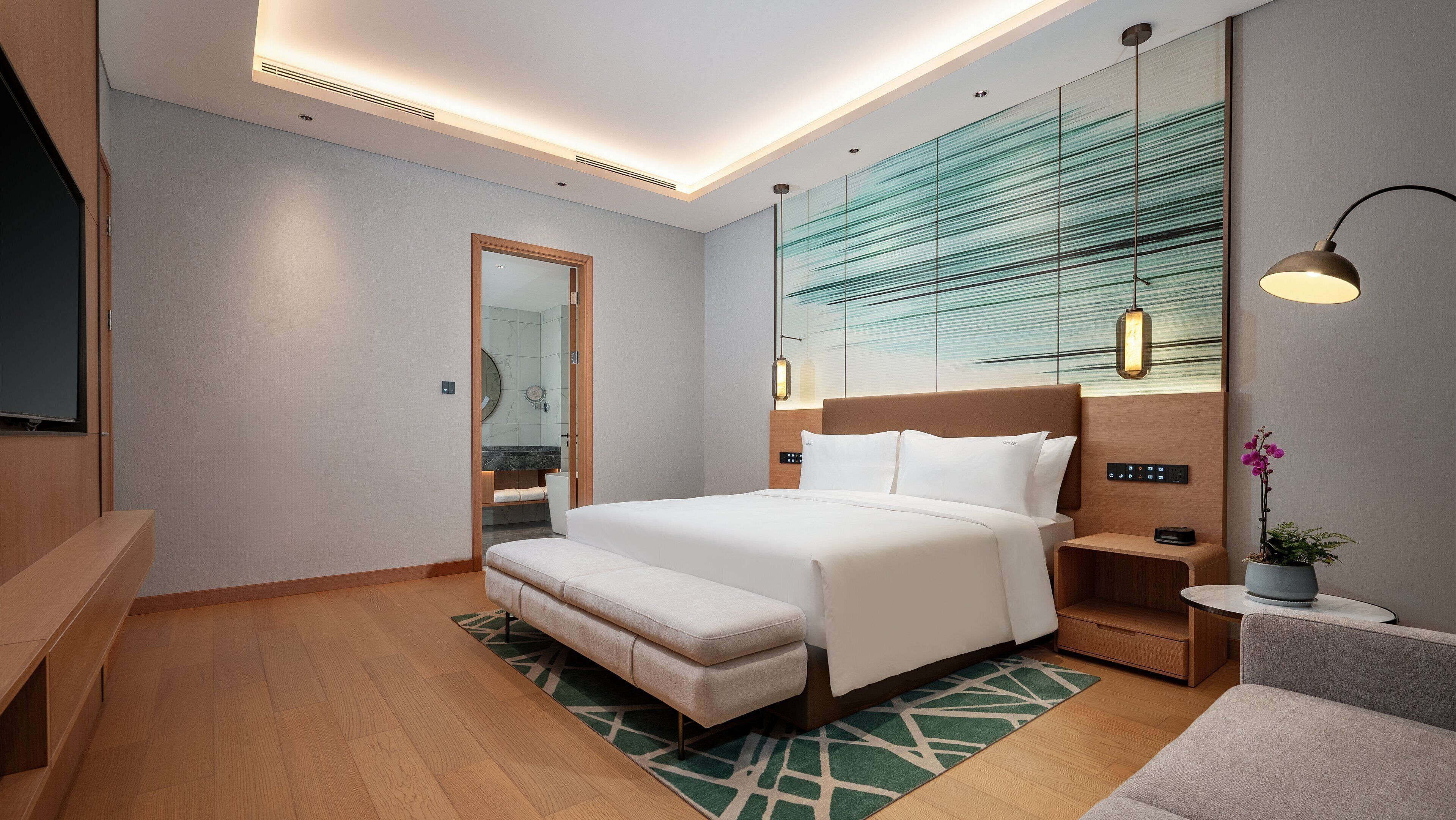 A modern hotel room with a large bed, a bench, a bedside table, a TV, and a bathroom visible through an open door.