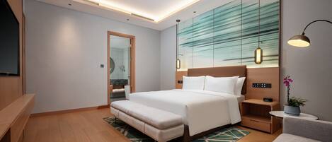 A modern hotel room with a large bed, a bench, a bedside table, a TV, and a bathroom visible through an open door.