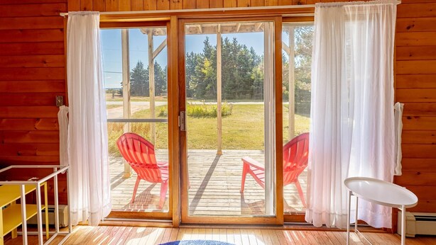 Una cabaña de madera con un ventanal corredero que da a una terraza exterior con sillas rojas y una mesa blanca.