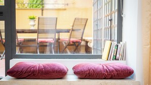 Ein Fenster mit zwei Kissen, eine Bücherwand voll Bücher und Blick auf eine Terrasse mit Stühlen und einem Tisch.