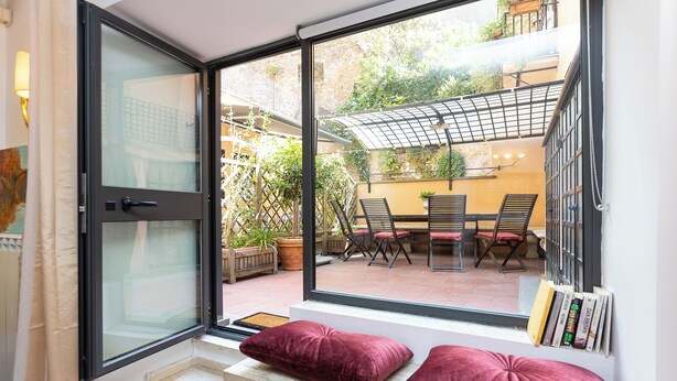 Ein Zimmer mit großem Fenster, das auf eine Terrasse mit Tisch und Stühlen, ein Bücherregal und eine gepolsterte Bank blickt.