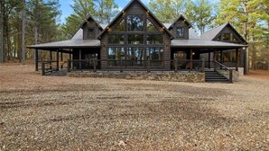 A log cabin with a large central window, a covered porch, and a stone foundation. - Hot Tub · Detached Game Rm · Pool Table | Dogs OK (Broken Bow)