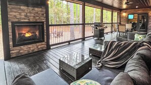 A cozy living room with a fireplace, large windows, and a deck area with outdoor furniture. - Hot Tub · Detached Game Rm · Pool Table | Dogs OK (Broken Bow)