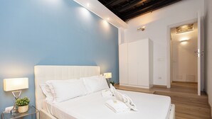 A neatly made bed with white linens and pillows, a bedside table with a lamp and a small plant, and a wardrobe in the background. - Rental in Rome Roma Campus Martius Apartment (Rome)