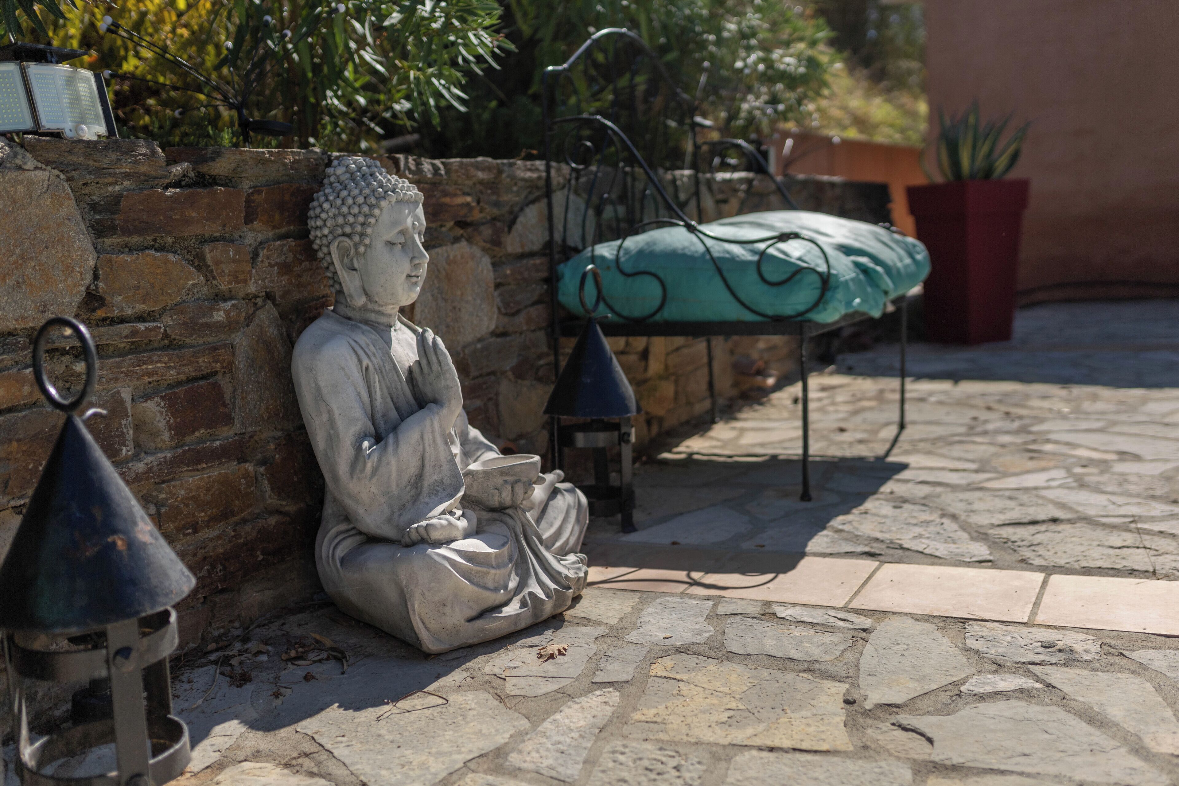 Une statue de Bouddha est assise sur un sol en pierre, à côté d’un mur de pierre. En arrière-plan, on aperçoit un banc en métal et une plante en pot.