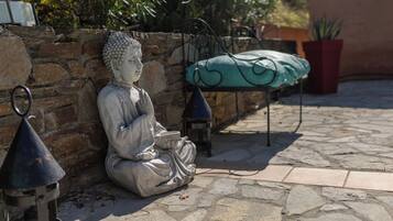 Une statue de Bouddha est assise sur un sol en pierre, à côté d’un mur de pierre. En arrière-plan, on aperçoit un banc en métal et une plante en pot.