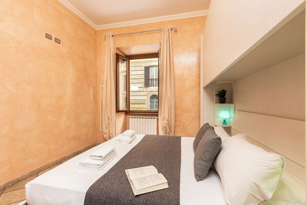 Habitación de hotel con una cama, una mesita de noche con lámpara, una ventana con cortinas y vistas a un edificio.